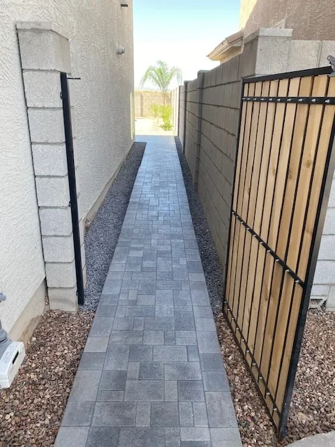 Gray paver walkway with river rock borders and black metal gate in side yard